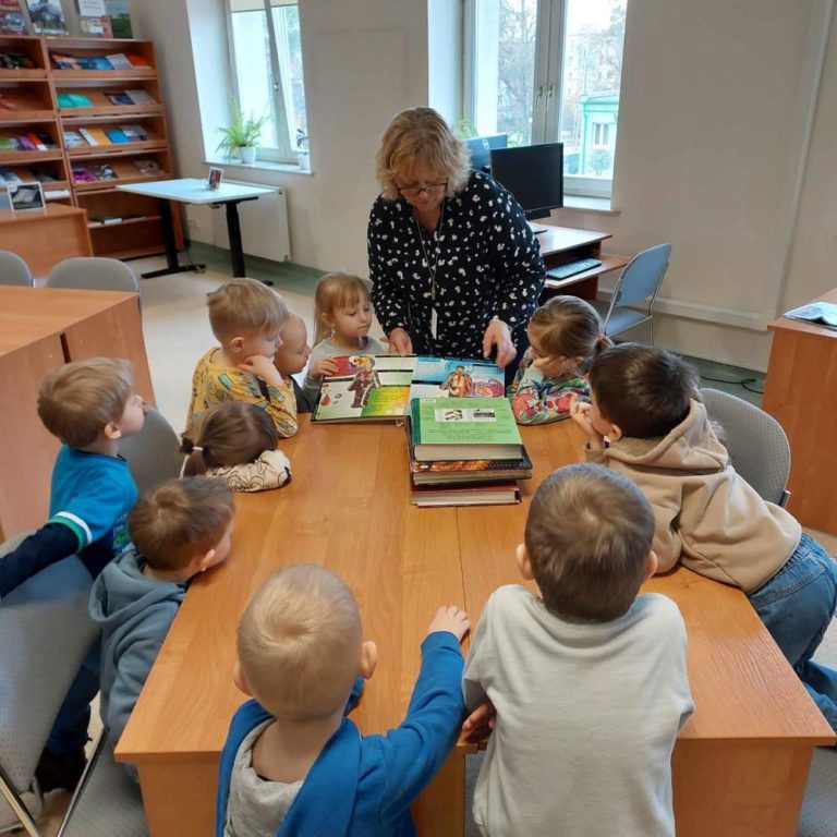 Grupa przedszkolaków podczas wizyty w bibliotece ogląda wydawnictwa książkowe dla dzieci