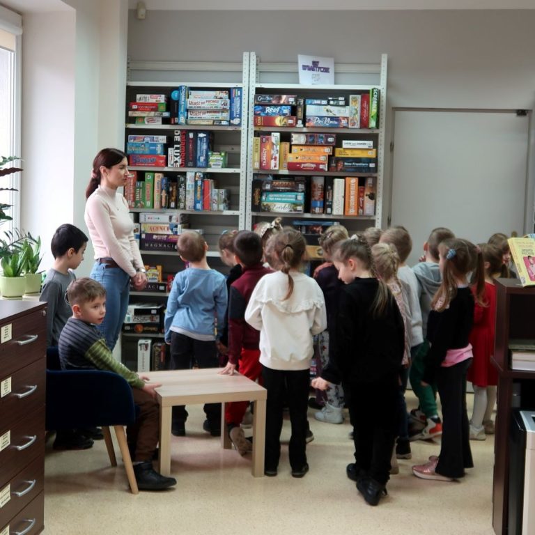 Bibliotekarka pokazuje dzieciom gry dydaktyczne zgromadzone w regałach