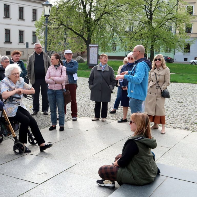 Grupa ludzi w różnym wieku zebranych wokół przewodnika w tle zabytkowe kamieniczki